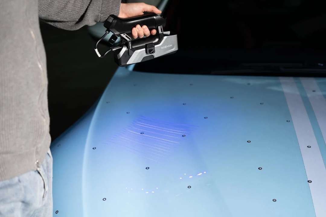 El escáner 3D de mano proyecta líneas láser azules paralelas sobre el capó de un automóvil cubierto de marcadores de referencia negros