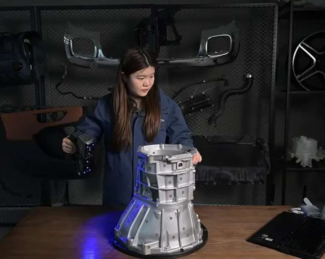 Un técnico maneja un escáner 3D de mano con láser azul; se escanea una gran carcasa de aluminio de una caja de cambios colocada sobre una mesa giratoria