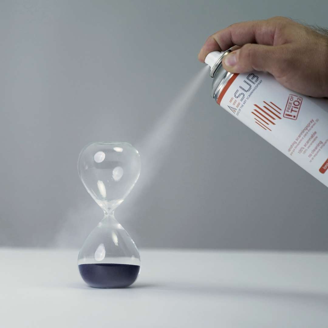 Mano aplicando AESUB Orange sobre un reloj de arena de vidrio transparente desde una distancia de aproximadamente 15–20 cm, niebla blanca fina visible