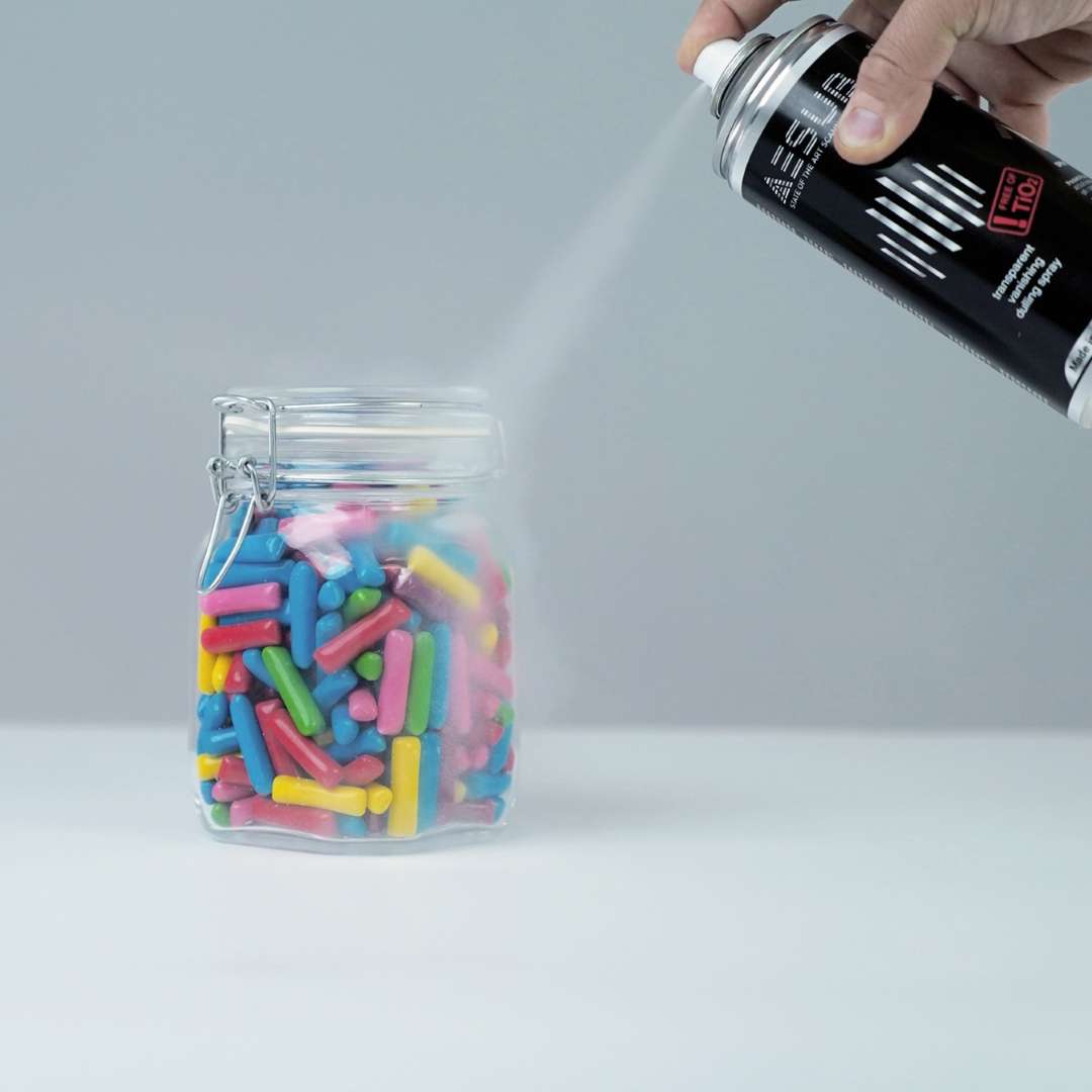 Mano aplicando AESUB Transparent sobre un tarro de vidrio con caramelos de colores, demostrando el ángulo de spray y la distancia de trabajo correctos