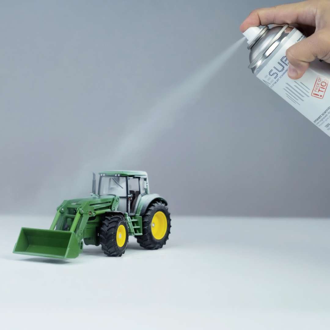 Mano aplicando AESUB White sobre un modelo de tractor verde a la distancia correcta, recubriendo la superficie con una fina niebla blanca