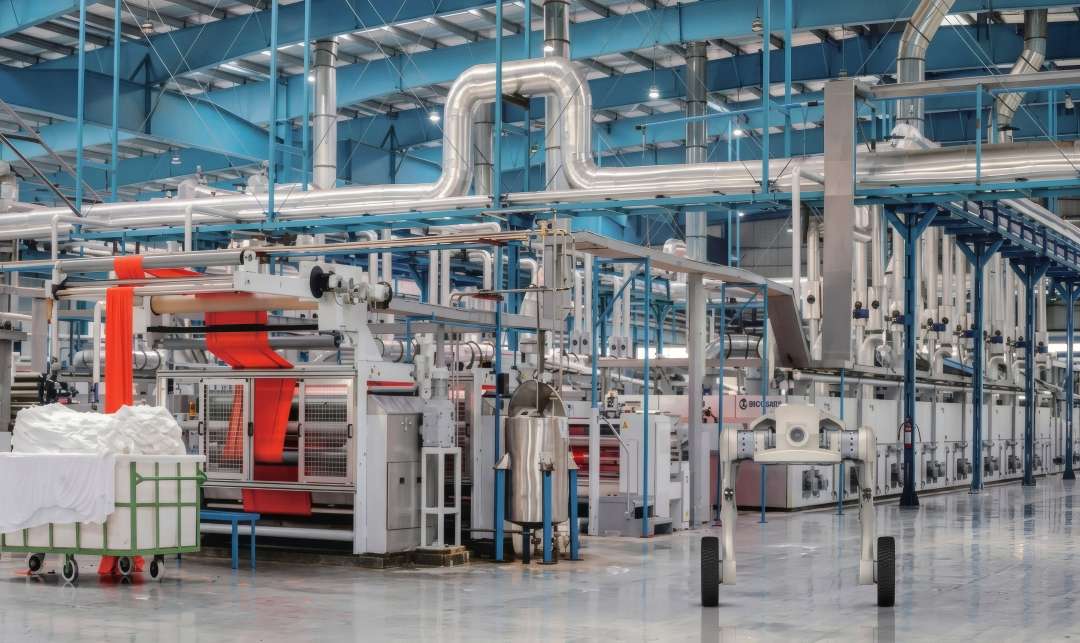 A bright factory workshop with silver pipes under the ceiling, a red fabric processing line, a trolley with white cloth and a white structure on two wheels in the foreground.