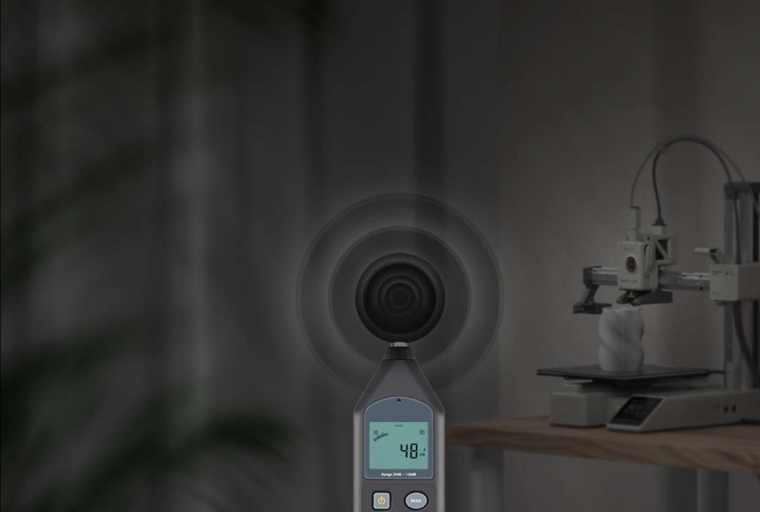 Sonómetro con pantalla digital en primer plano; detrás, sobre una mesa, una impresora 3D imprime un modelo blanco retorcido.