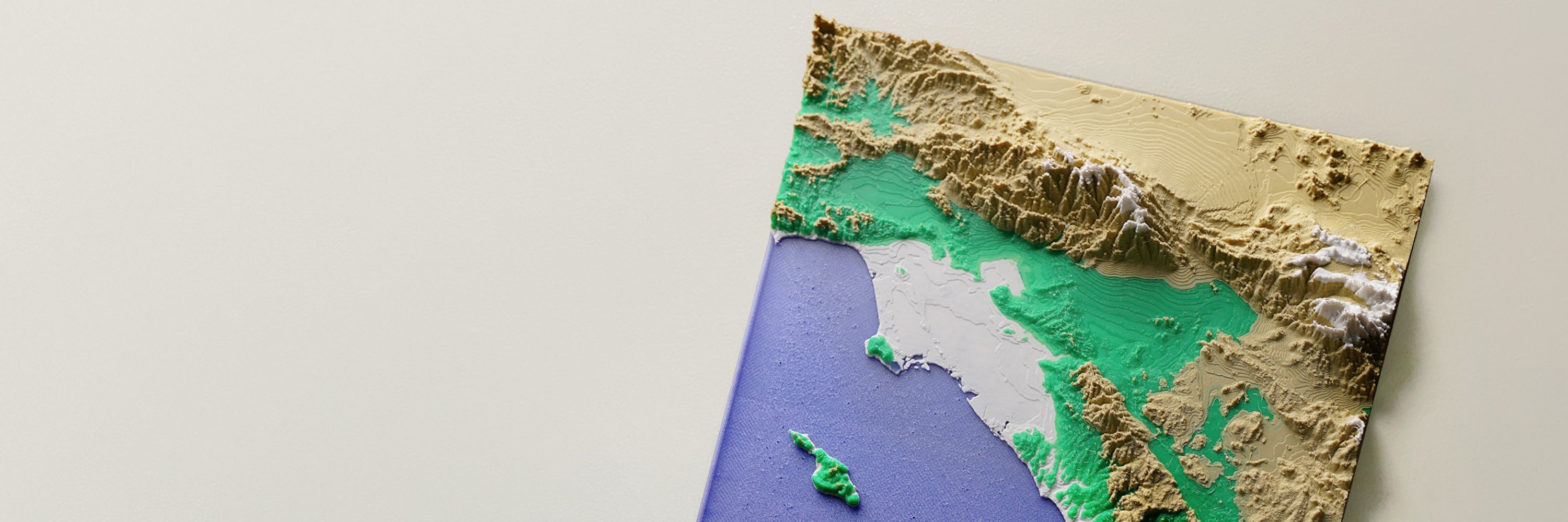 Mapa topográfico en relieve de una costa con cordilleras de color arena, cumbres blancas, valles verdes, un mar azul violáceo y una pequeña isla.
