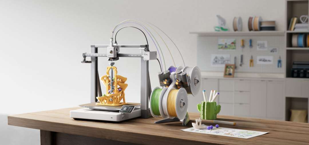 Una impresora de sobremesa con una pequeña pantalla en la base imprime un modelo amarillo con bolitas moradas; a la derecha, en un soporte, hay bobinas verde, amarilla y blancas.