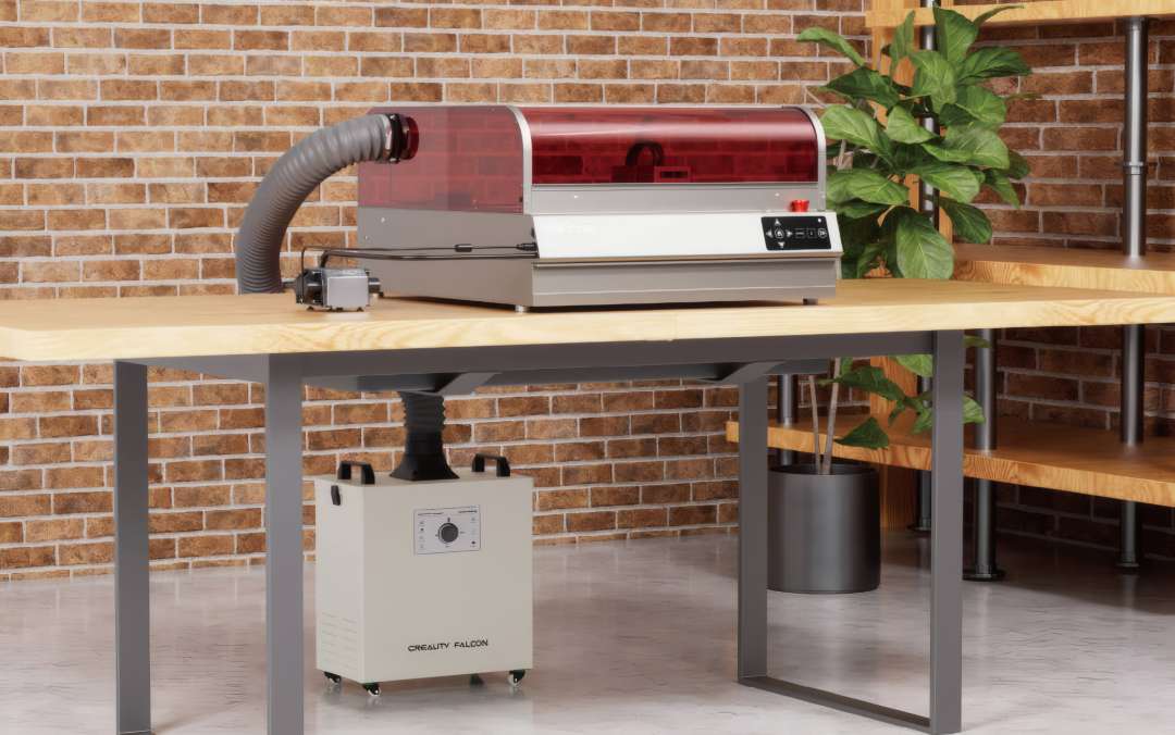 Grabadora láser Creality Falcon2 Pro S 22W sobre banco de trabajo de madera en estudio con pared de ladrillo, manguera de extracción conectando la máquina a un purificador de humos Creality Falcon situado en el suelo bajo la mesa