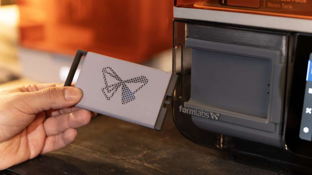 Una mano sostiene un panel gris extraíble con un logotipo de mariposa perforado junto al compartimento frontal abierto de una impresora Formlabs.