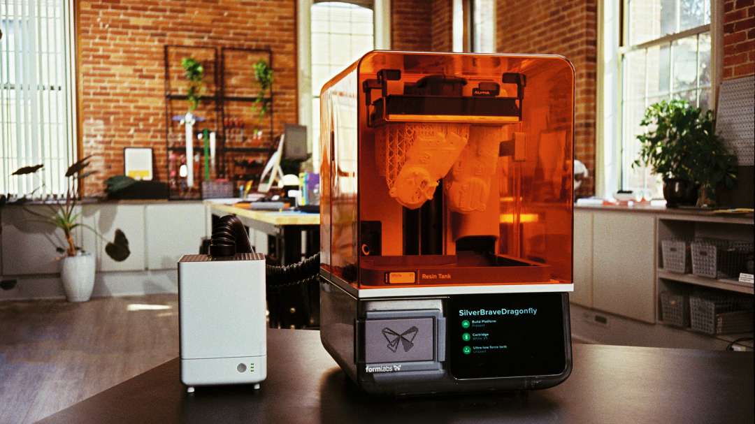 Impresora 3D de resina con tapa protectora naranja y piezas sobre soportes reticulares; al lado, una unidad blanca con una manguera conectada.