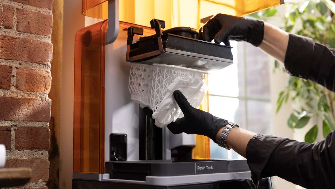 Unas manos con guantes extraen la plataforma de impresión con una pieza impresa blanca sobre soportes reticulares por encima del depósito de resina.