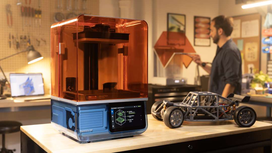 Impresora 3D de resina con tapa naranja tintada y pantalla táctil junto a un chasis gris de buggy impreso en un taller.