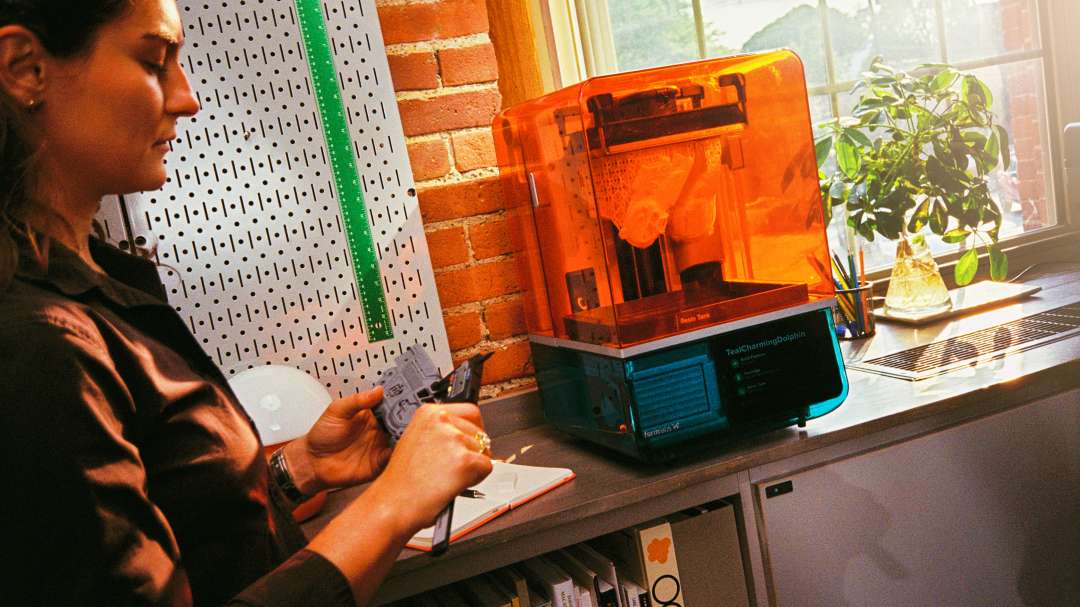 En un estudio hay una impresora 3D de resina de sobremesa con tapa naranja translúcida, y una persona examina una pieza gris.