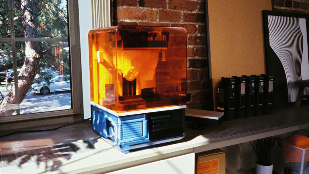 Impresora 3D de resina de sobremesa con tapa naranja translúcida y base azul junto a cartuchos negros en una estantería.