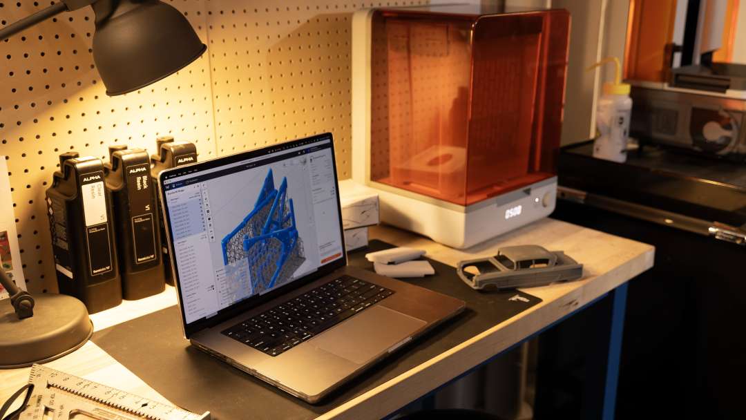 Impresora con tapa naranja junto a un portátil cuya pantalla muestra la preparación de soportes, y un modelo de coche gris terminado