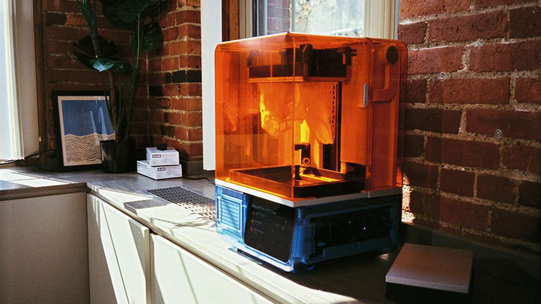 Impresora 3D de resina cerrada con tapa naranja semitransparente y pantalla en la parte delantera sobre una encimera junto a una pared de ladrillo