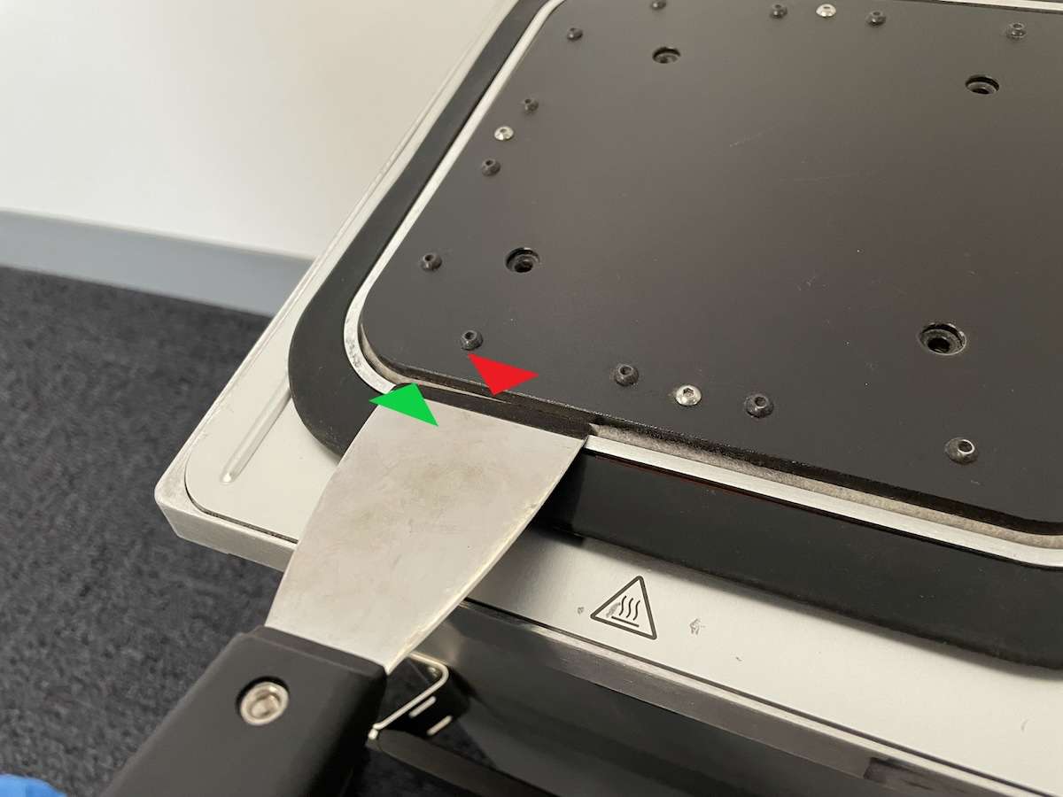 Espátula (flecha verde) introducida en el espacio entre la parte inferior de la plataforma de impresión y el sello de fieltro de la cámara Fuse 1 para levantarla suavemente