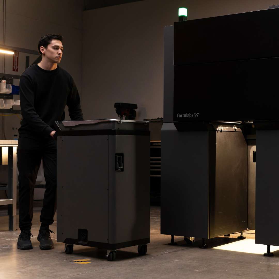 A technician wheels a mobile cart towards an open Formlabs Fuse X1 industrial printer with an illuminated build unit