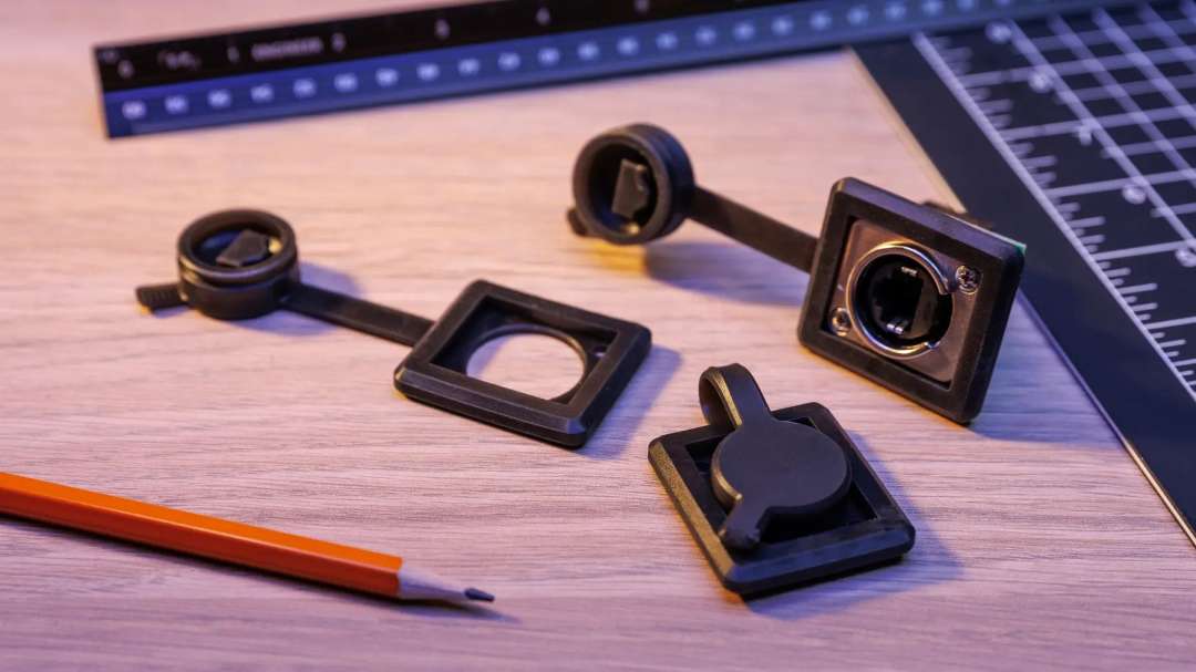 Sobre una mesa de madera, junto a una regla y un lápiz, hay marcos flexibles negros con tapones sujetos por correas, y uno de ellos está colocado en una toma de conector.