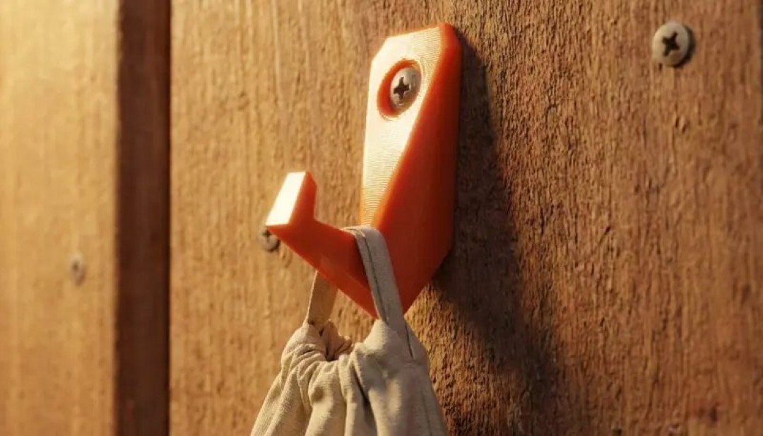 Gancho naranja creado mediante impresión 3D que sostiene una bolsa en una pared de madera.