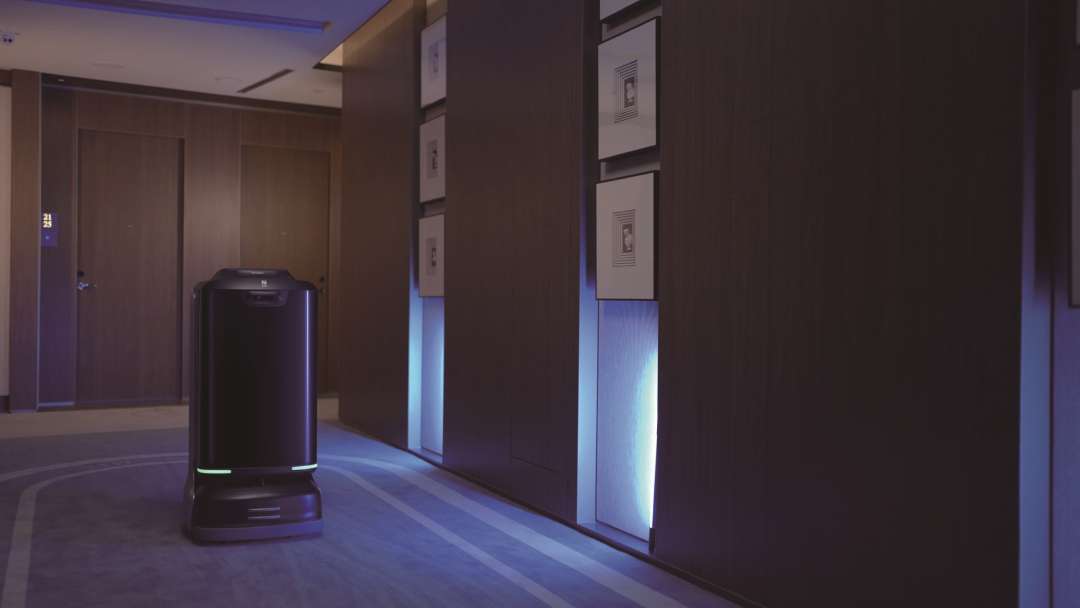 Robot de entrega PUDU FlashBot aproximándose al banco de ascensores en un pasillo de hotel con poca luz, con la tira indicadora azul-verde iluminada a nivel del suelo