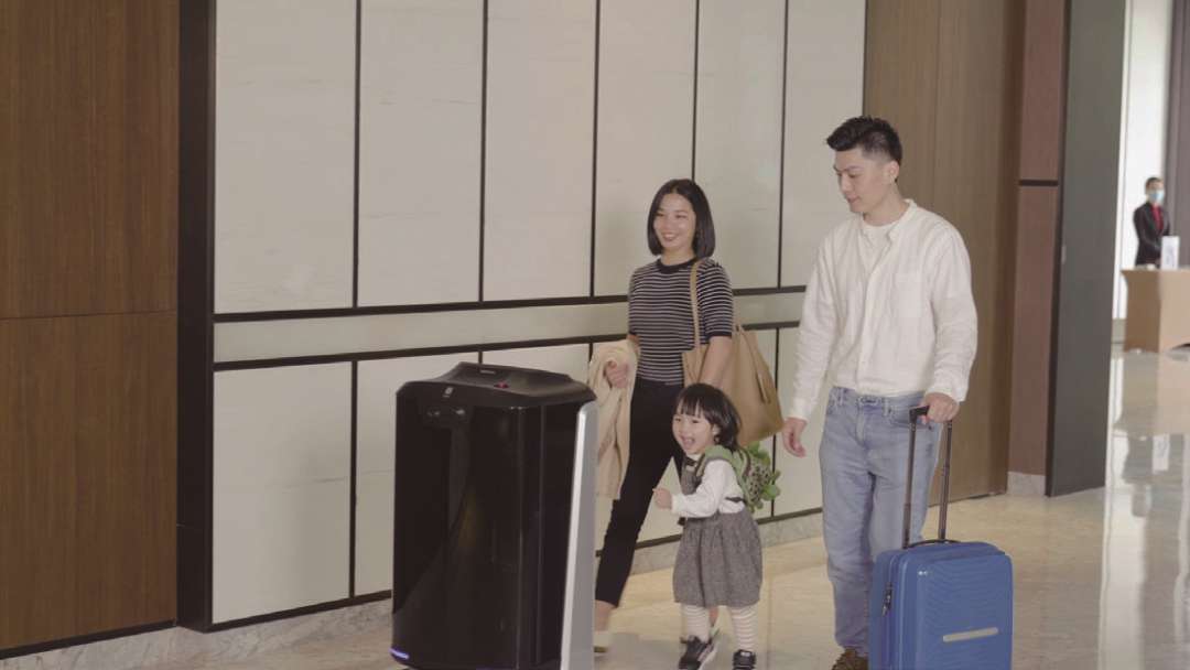 Robot de entrega PUDU FlashBot pasando junto a una familia de tres personas — madre, padre con maleta e hijo pequeño — en el iluminado vestíbulo de un hotel durante el check-in