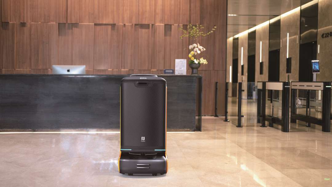 Robot de entrega PUDU FlashBot situado frente al mostrador de recepción de un hotel moderno de alto nivel, con panelado de madera cálida e iluminación decorativa