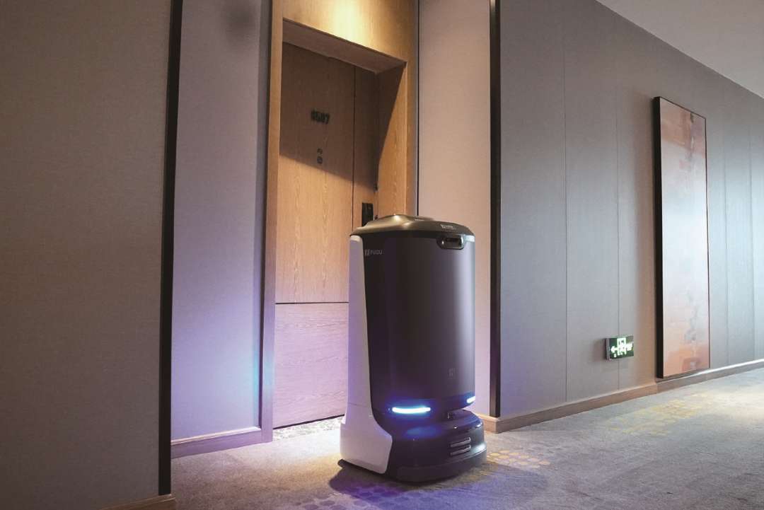 Robot de entrega PUDU FlashBot esperando ante la puerta numerada de una habitación de hotel en un pasillo enmoquetado, con el aro indicador azul iluminado, listo para completar la entrega