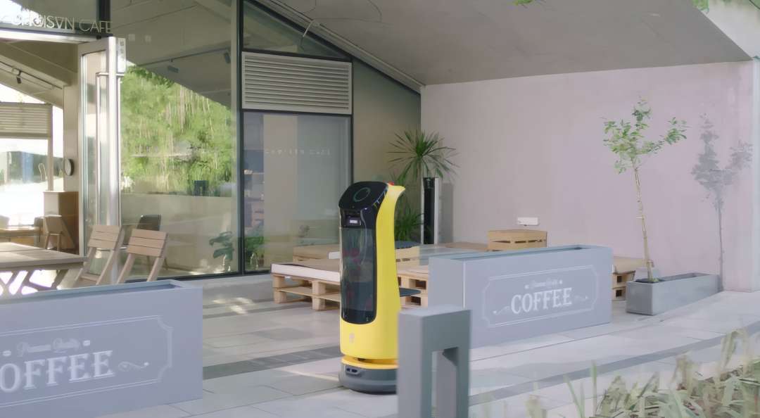 Жовтий робот PUDU KettyBot Pro, що автономно рухається відкритою терасою кафе між вазонами і зонами для сидіння без напрямних рейок