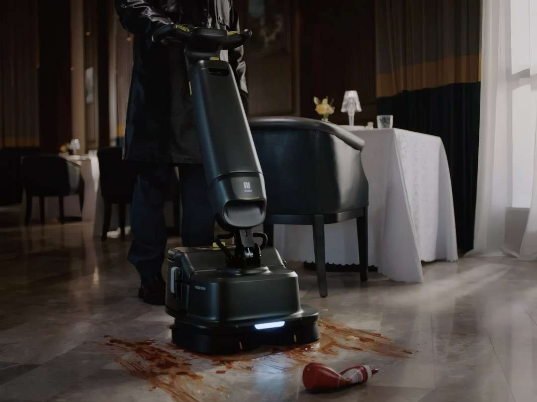 Operario con uniforme oscuro usando la fregadora PUDU SH1 para limpiar un derrame de kétchup sobre suelo de mármol en comedor de restaurante de lujo