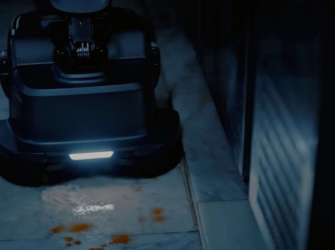 Chasis de la fregadora PUDU SH1 iluminando un derrame en borde de pared con su tira LED frontal en un servicio de cocina comercial oscuro
