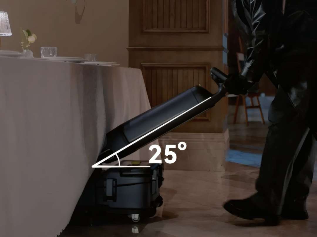 Fregadora PUDU SH1 inclinada a 25 grados para limpiar bajo una mesa de comedor cubierta con mantel, con solución de limpieza visible en el suelo