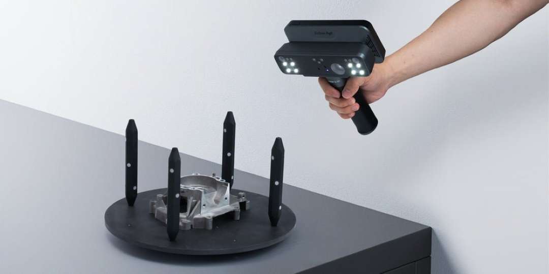 Una mano sostiene un escáner de mano negro sobre una pieza metálica fundida situada en una plataforma redonda, en cuyos bordes hay cuatro soportes oscuros con puntos blancos.