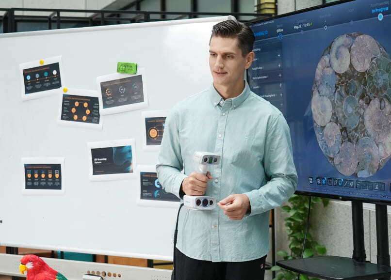 Un hombre muestra un escáner 3D portátil frente a una pizarra blanca y una pantalla digital